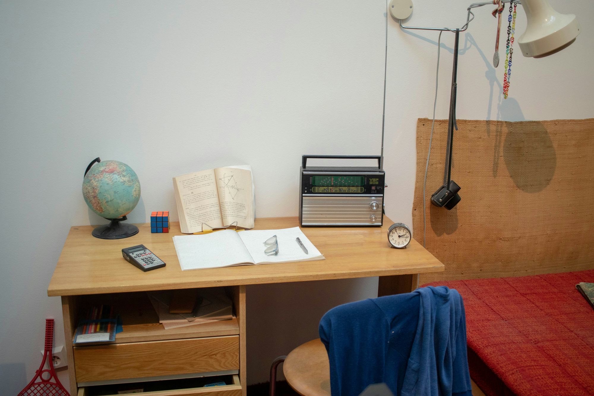 A student's desk: a globe, a Rubik's cube, an open textbook, a ruled notebook, a radio and an alarm clock under an angled lamp.